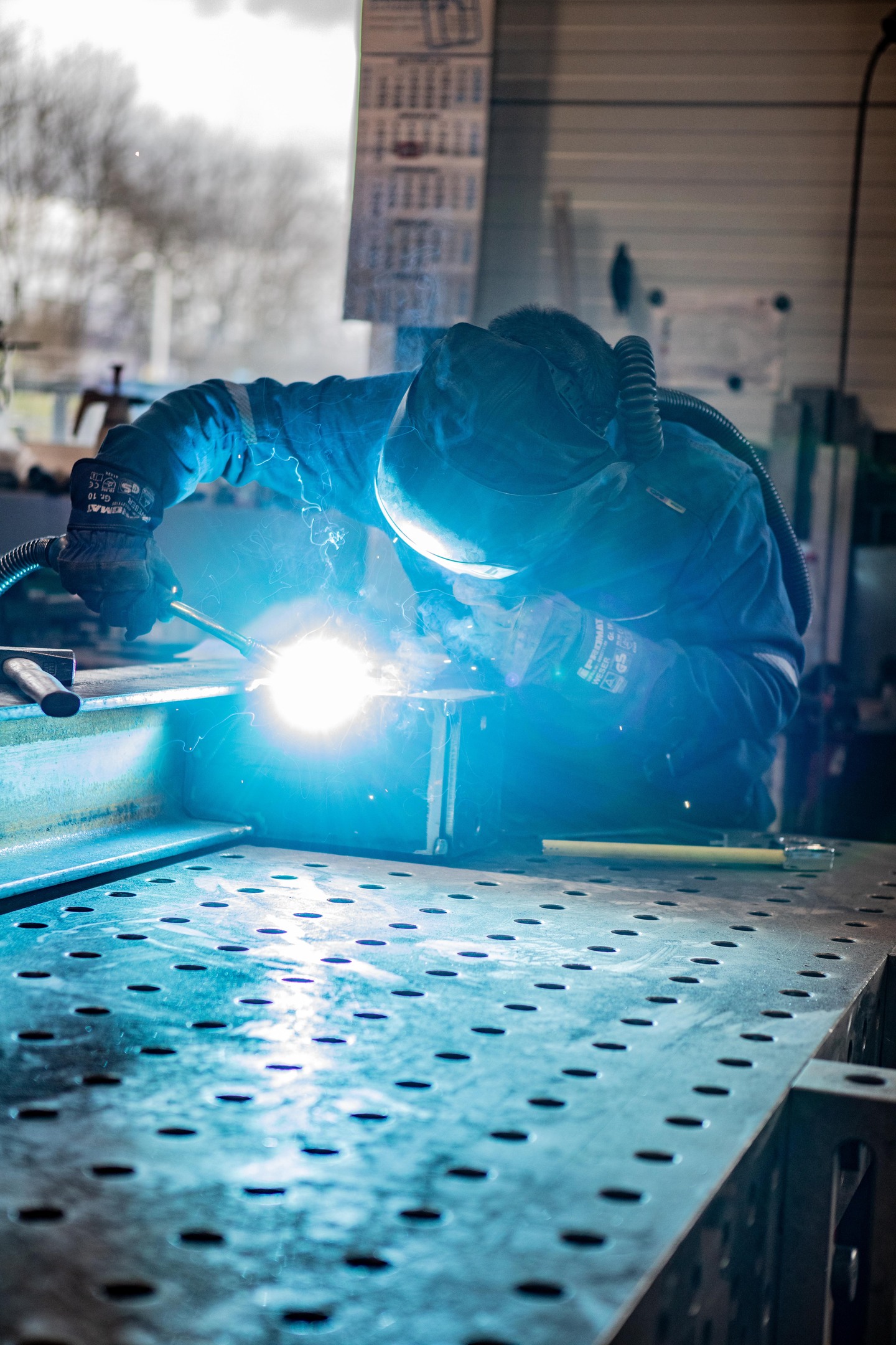 Ein Schweißer in Schutzkleidung und Helm schweißt Metallteile in einer Werkstatt.