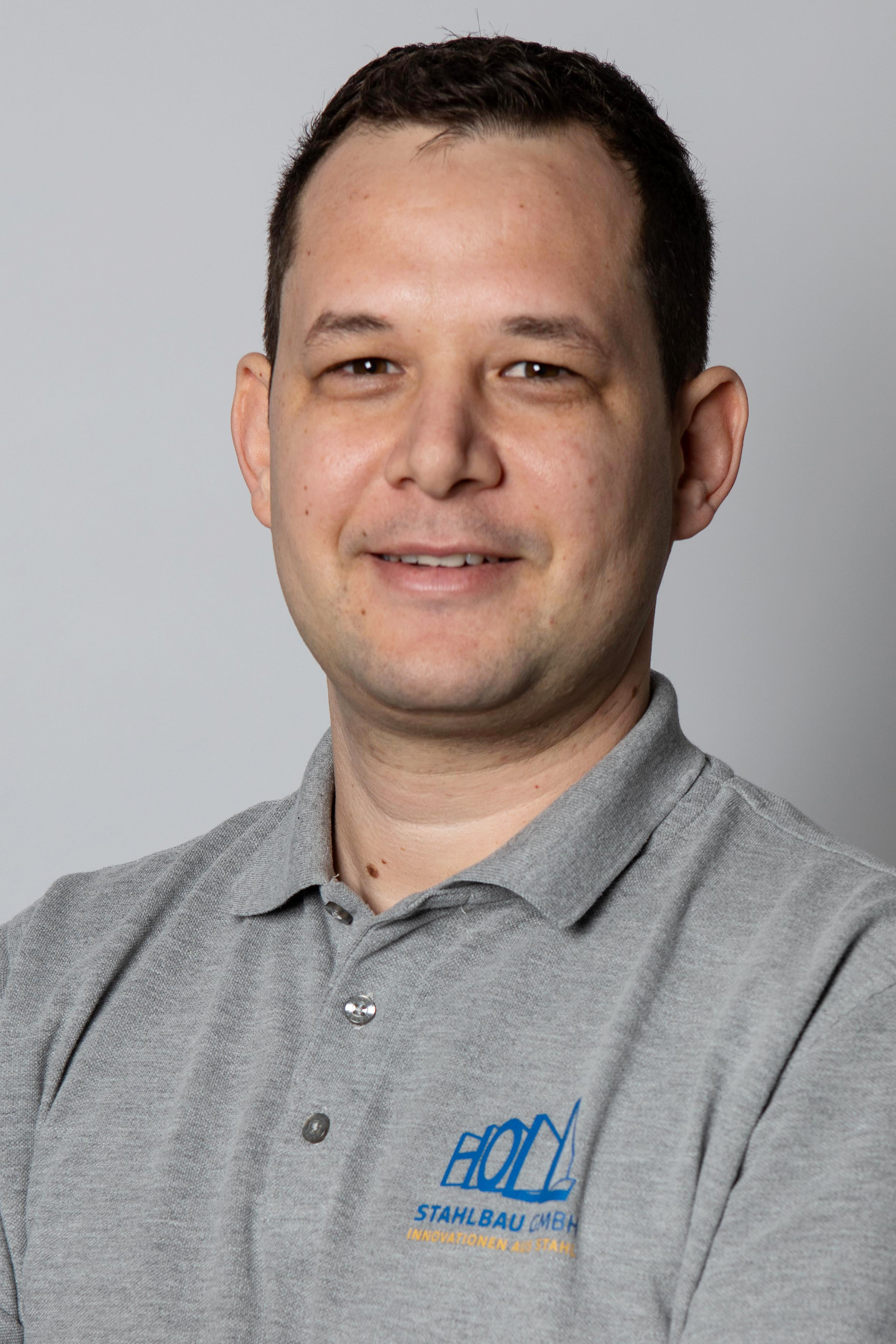 Ein Mann mit kurzem dunklem Haar trägt ein graues Poloshirt mit dem Logo "HOLD Stahlbau GmbH" und einem blauen Schriftzug.