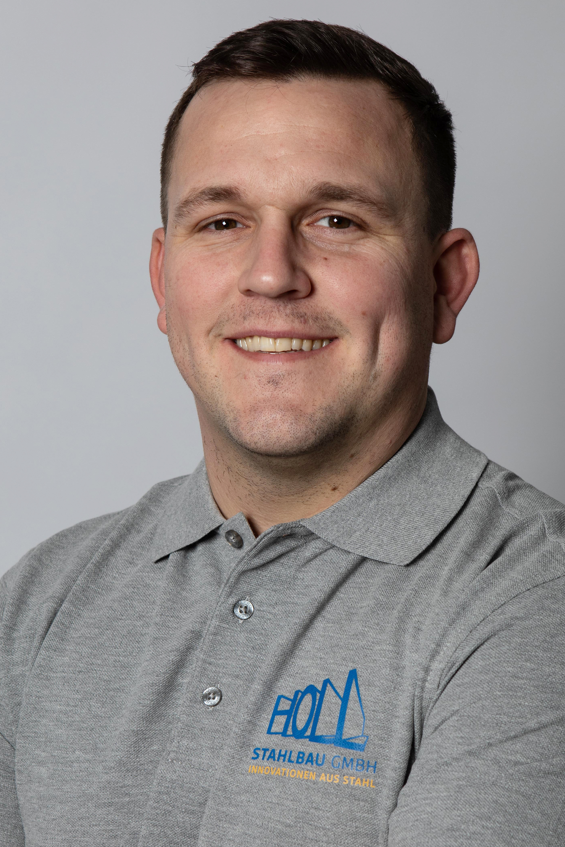 Ein Mann mit kurzem Haar und lächelndem Gesicht trägt ein graues Poloshirt mit dem Logo und dem Text „HOLM STAHLBAU GMBH - INNOVATIONEN AUS STAHL“.