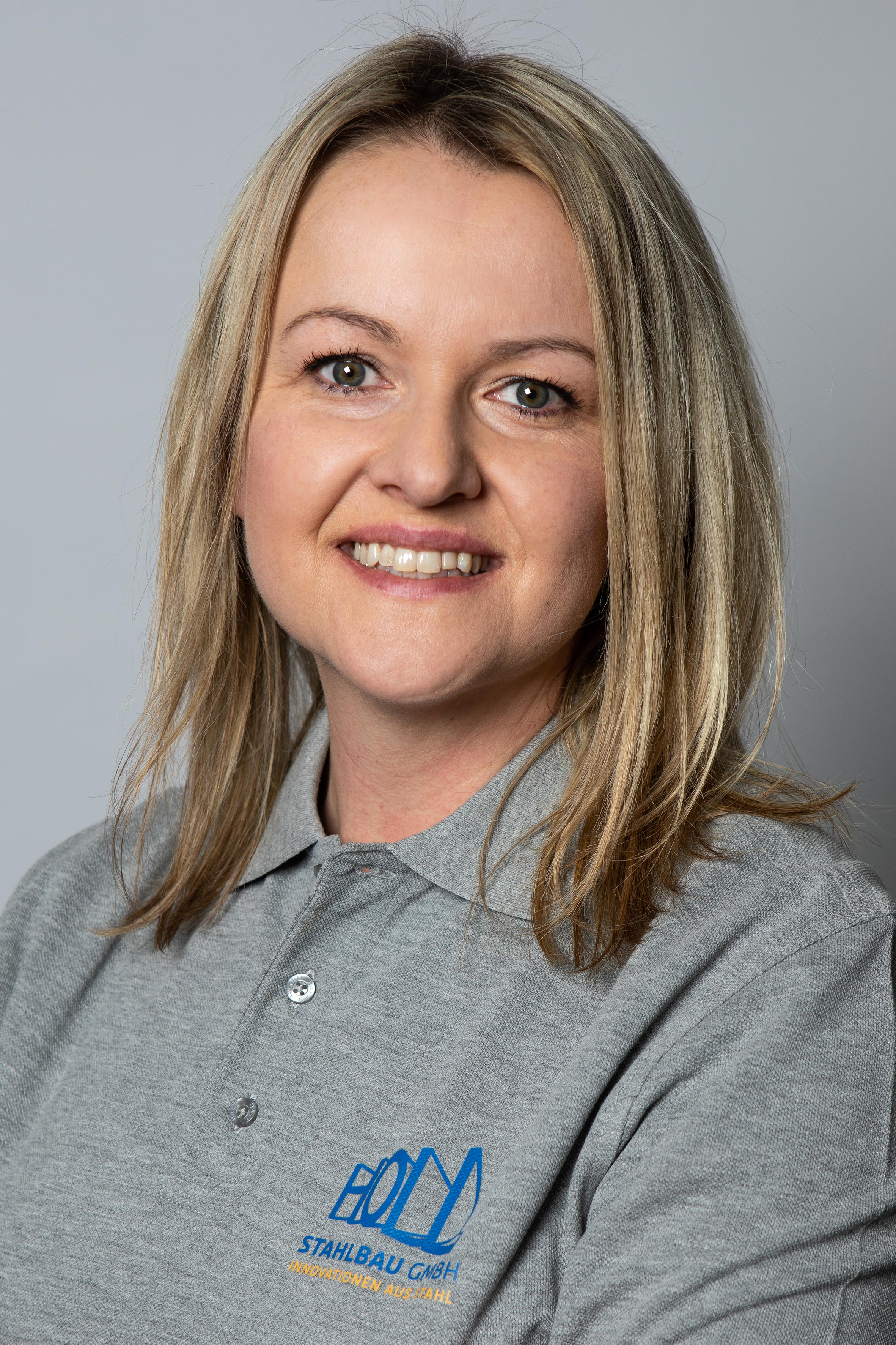 Eine Person mit blonden Haaren trägt ein graues Poloshirt mit dem Logo und dem Text "STAHLSBAU GMBH" und lächelt in die Kamera.