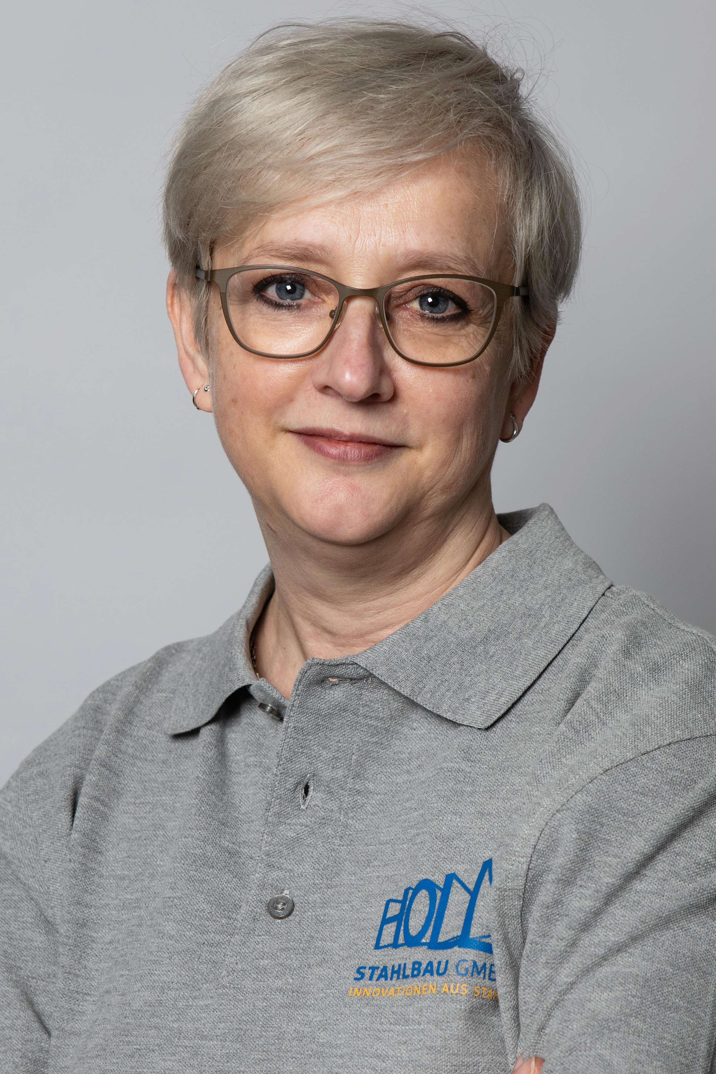 Eine Person mit kurzen blonden Haaren, Brille und Ohrringen trägt ein graues Poloshirt mit dem Logo "BOLD Stahlbau GmbH" und dem Slogan "Innovationen aus Stahl".