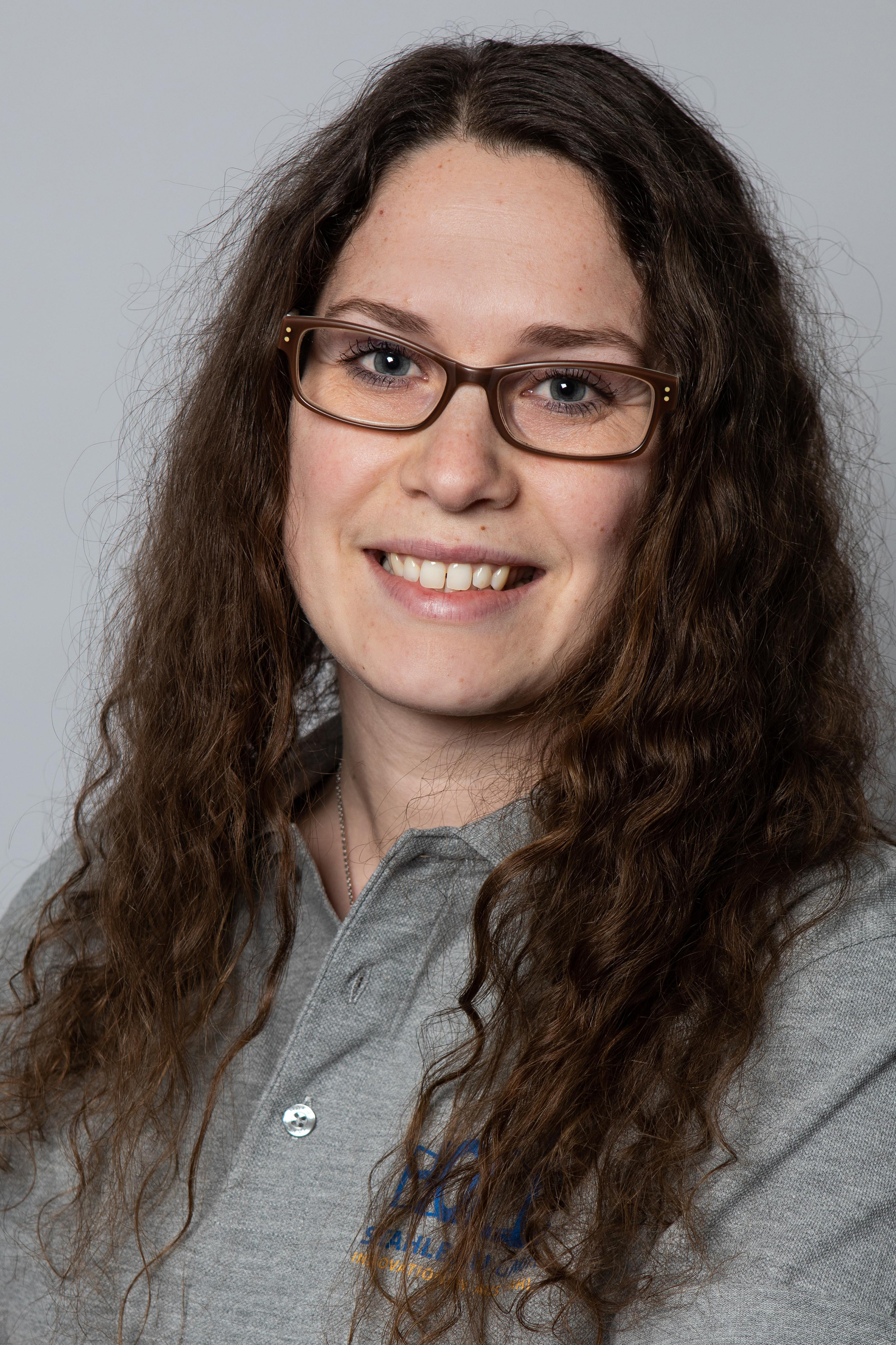 Eine Person mit langen, welligen Haaren und Brille lächelt in die Kamera, während sie ein graues Hemd trägt.