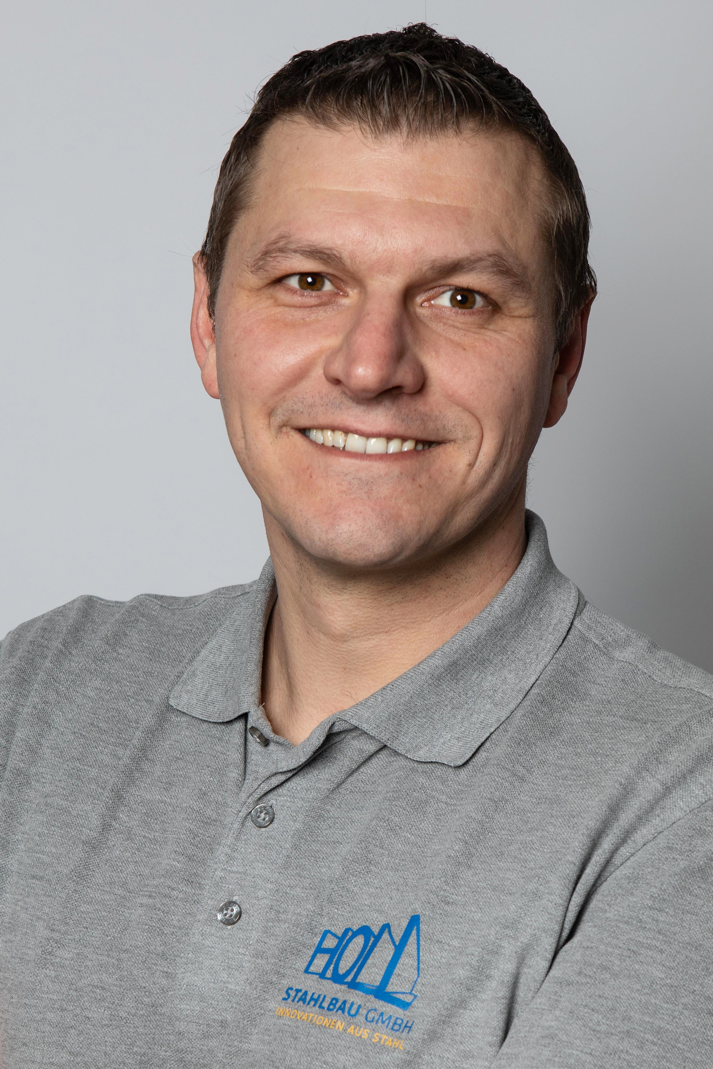 Ein Mann mit kurzen dunklen Haaren und einem grauen Poloshirt mit dem Logo "Stahlbau GmbH" lächelt in die Kamera.