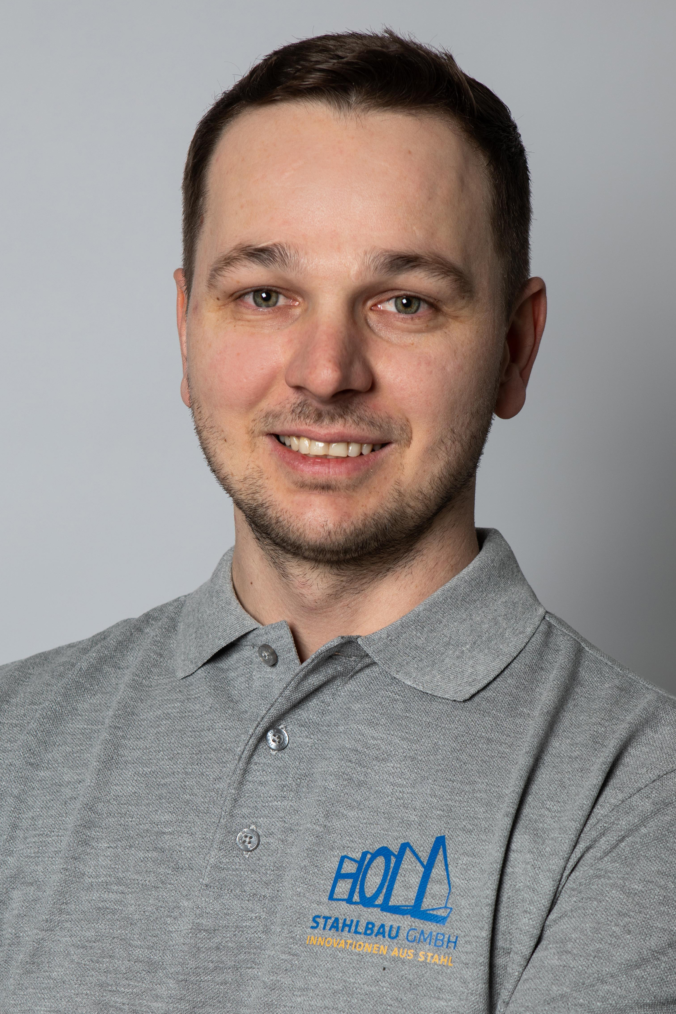 Ein Mann mit kurzem Haar und Bart trägt ein graues Poloshirt mit dem Logo und dem Text "HOLZ STahlbau GmbH - Innovationen aus Stahl".