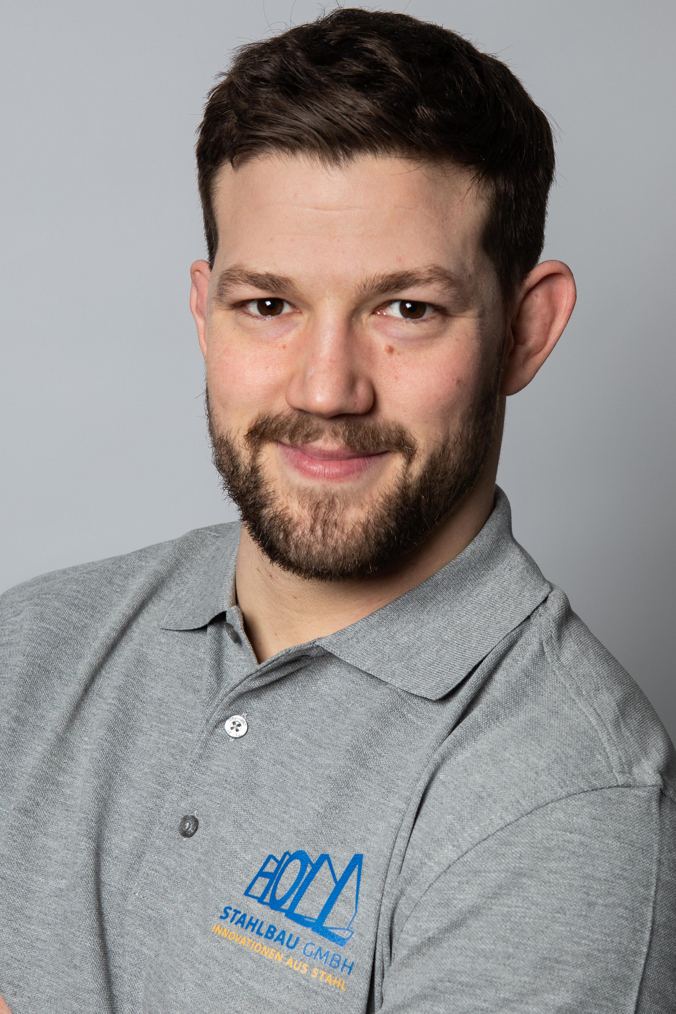 Ein Mann mit Bart und kurzen Haaren trägt ein graues Poloshirt mit dem Logo "HOLZ STAHLBau GMBH" und dem Slogan "Innovationen aus Stahl".